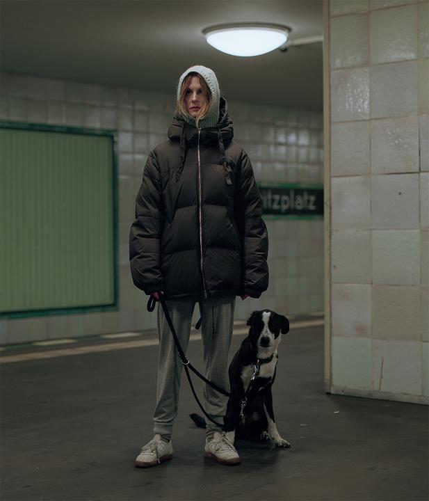A woman in a puffer jacket and a dog on a leash stand in a subway station.