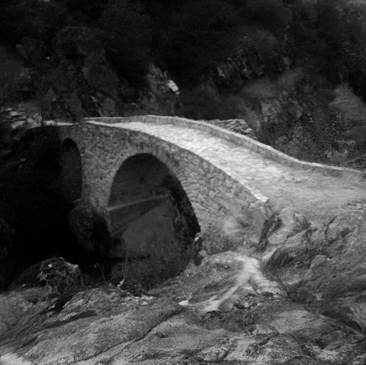 A stone arch bridge spans a rocky gorge with dark foliage in the background.