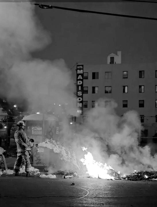 A firefighter sprays water on a large fire in the street at night, with the Hotel Madison sign visible in the background.