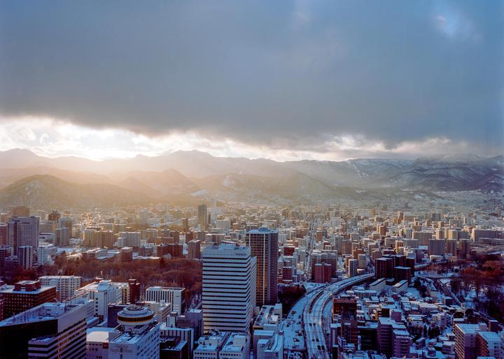 A city with many buildings and a highway is covered in snow, with mountains in the background under a cloudy sky.