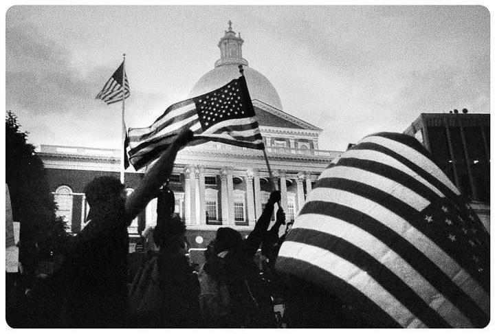 People hold American flags in front of a domed building with columns.