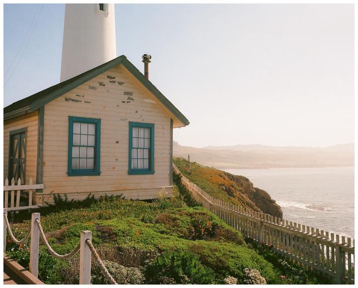 A small house with blue window frames sits on a cliff overlooking the ocean next to a lighthouse.