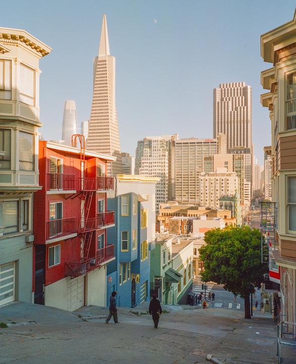 Two men walk down a steep street lined with colorful houses and tall buildings in the background.