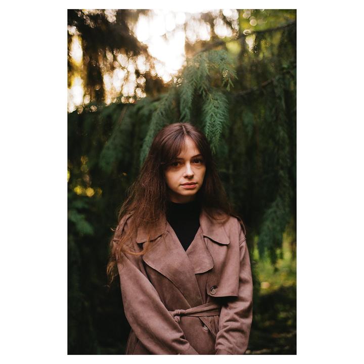 A woman with long hair and a trench coat stands in front of a dark green tree.
