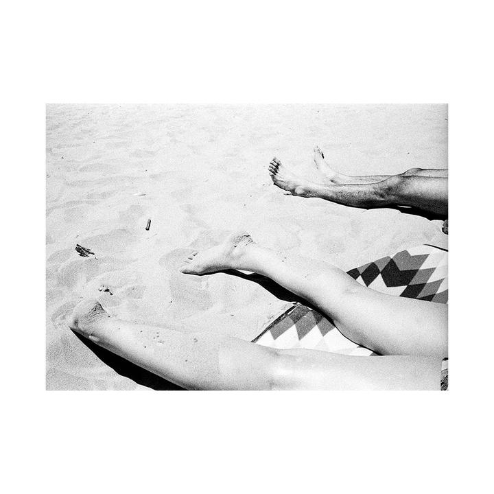 Two people are relaxing on a patterned blanket on a sandy beach.