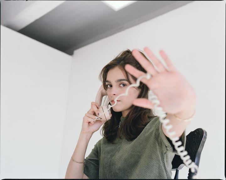 A woman holds a telephone receiver to her ear while extending her other hand toward the viewer, with the telephone cord in the foreground.