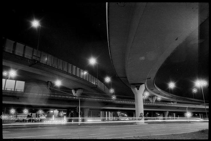 A multi-level highway interchange is illuminated by streetlights at night, with light trails from vehicles on the road below.