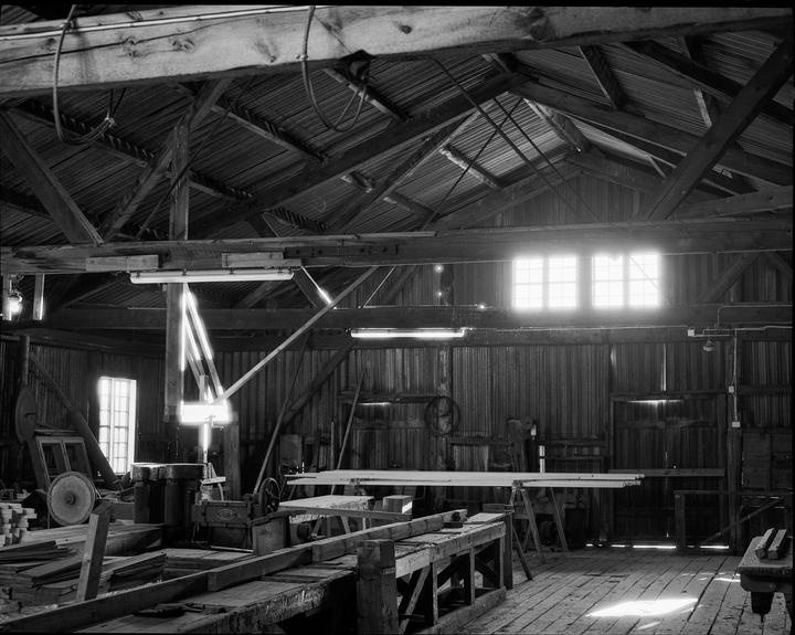 Sunlight streams through windows into a large, rustic wooden workshop filled with tools and lumber.