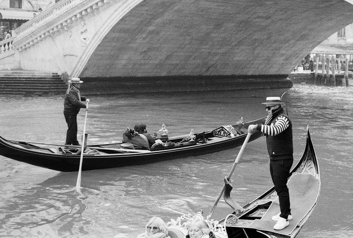 Two gondoliers in striped shirts and straw hats steer gondolas on a canal under a bridge, with passengers visible in the foreground and background.