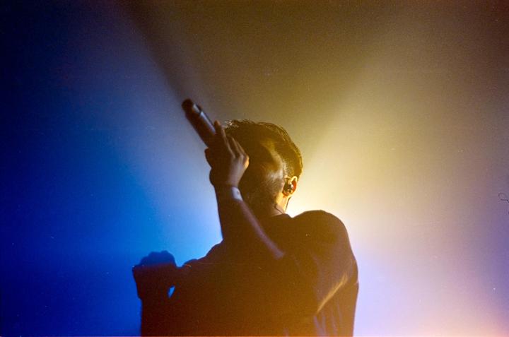 A man sings into a microphone on a stage with blue and yellow lighting.