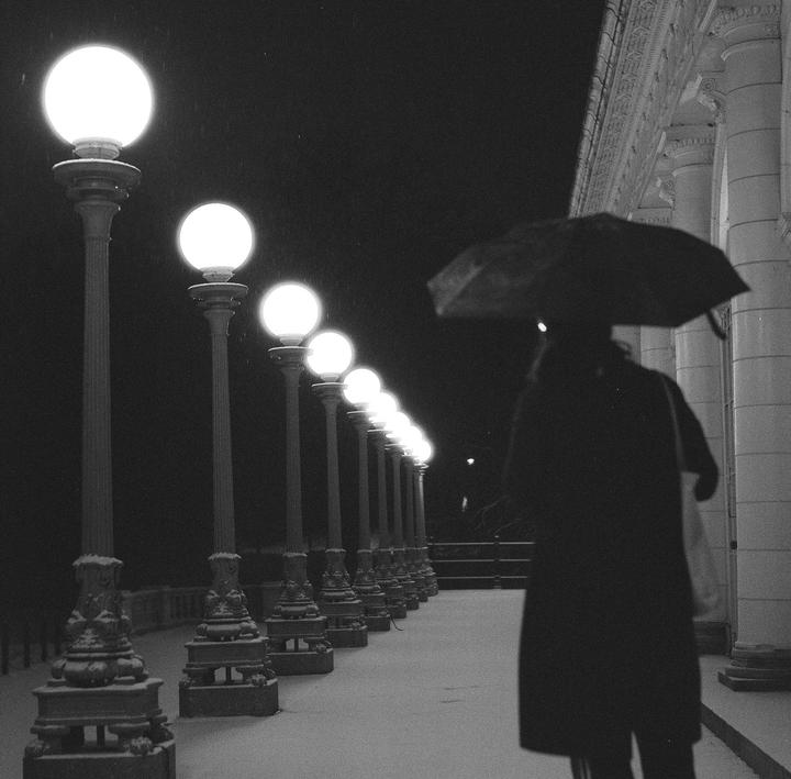 A person holding an umbrella walks past a row of glowing streetlights at night.
