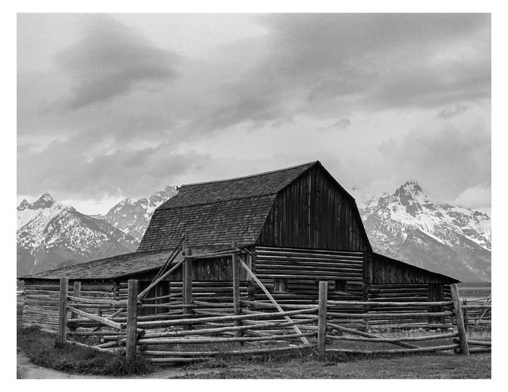 A wooden barn with a fence in front of it stands before snow-capped mountains under a cloudy sky.