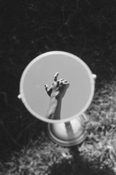 A hand is reflected in a round mirror, pointing upwards towards a bright sky.