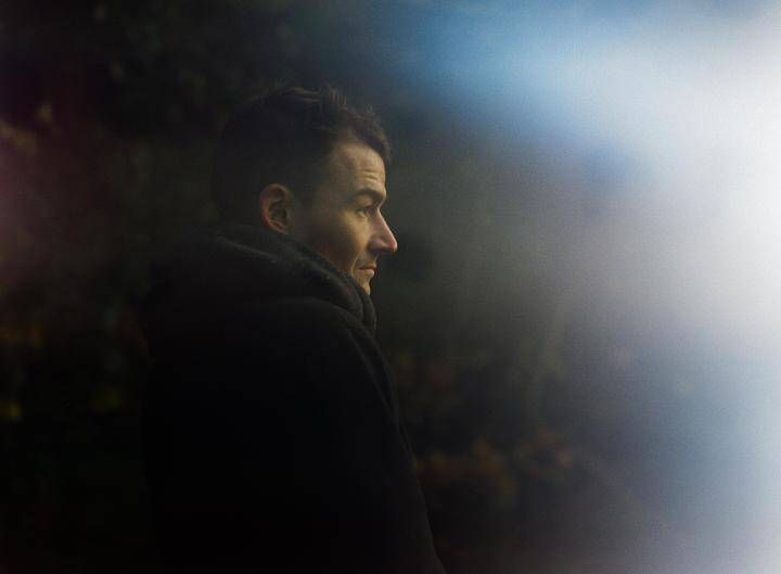 A man in a dark coat looks to the right with light on his face and a hazy background.