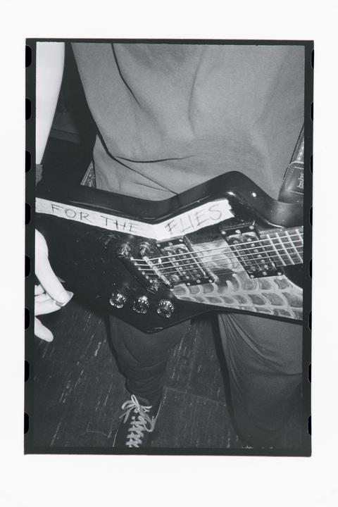 A person holds an electric guitar with "FOR THE FLIES" written on it.