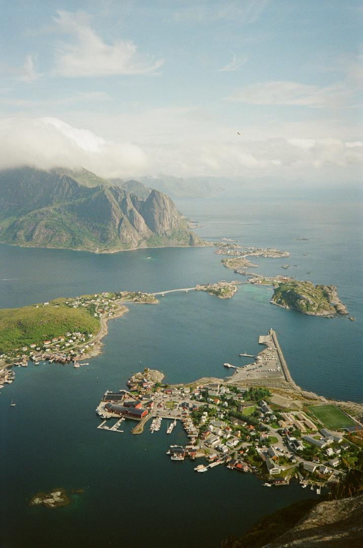 An aerial landscape shows a small coastal town with a harbor and a bridge connecting to other islands, surrounded by mountains and ocean.