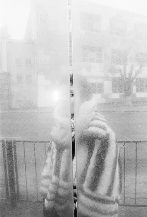 A person wearing a striped coat is reflected in a window, with a building and a tree visible in the background.