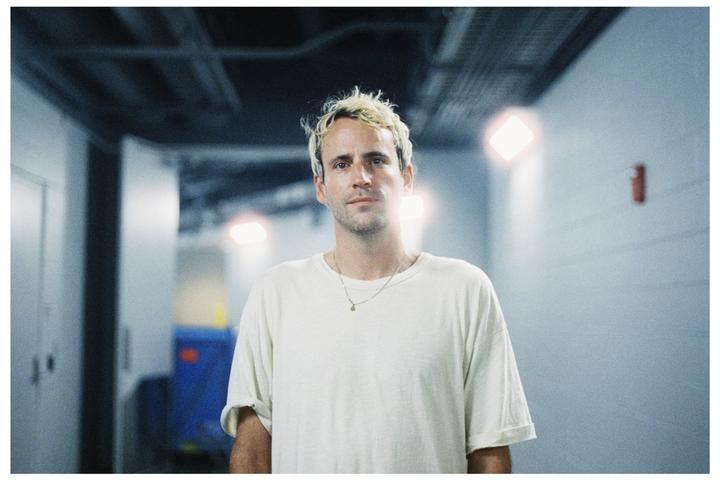 A man with bleached hair and a white t-shirt stands in a hallway with bright lights.