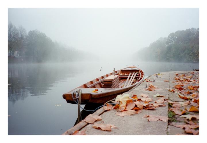 A wooden rowboat is moored to a concrete bank covered in autumn leaves, with a foggy lake and trees in the background.