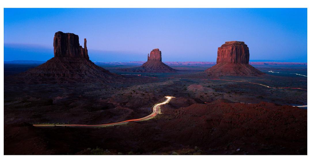 Three large rock formations stand in a desert landscape under a blue sky with light trails from vehicles on a winding road.