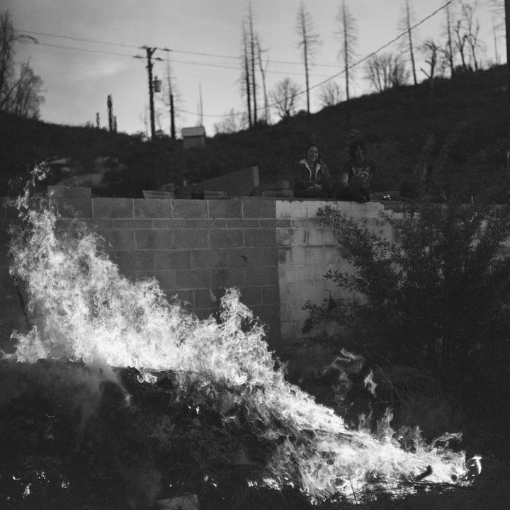 Two people sit on a concrete block wall above a large bonfire with a hillside and bare trees in the background.
