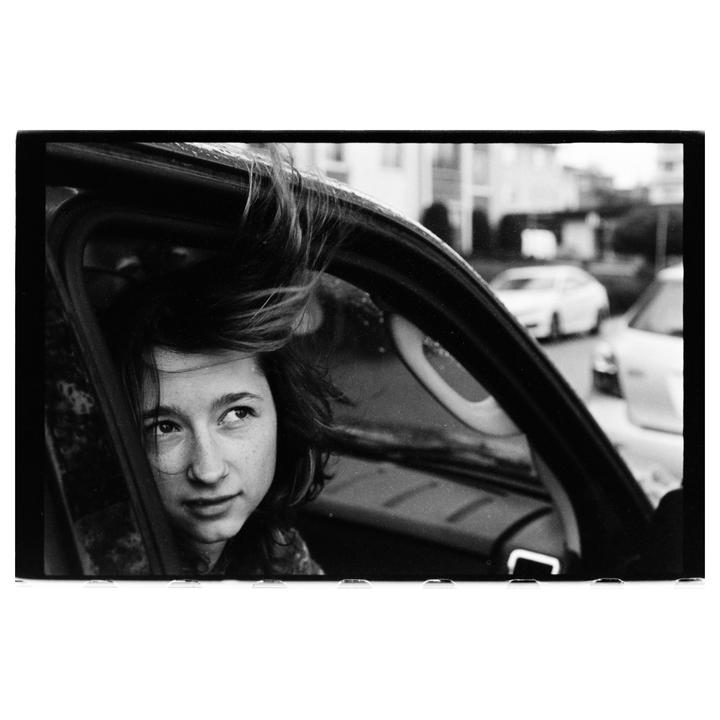 A girl with windblown hair looks out of a car window onto a street with other cars.