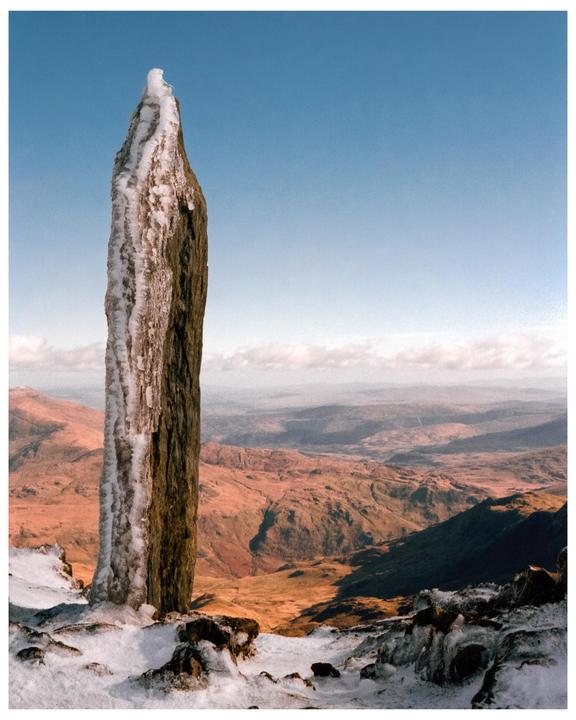 A tall, ice-covered stone pillar stands in a snowy landscape with mountains and a valley in the background under a clear sky.