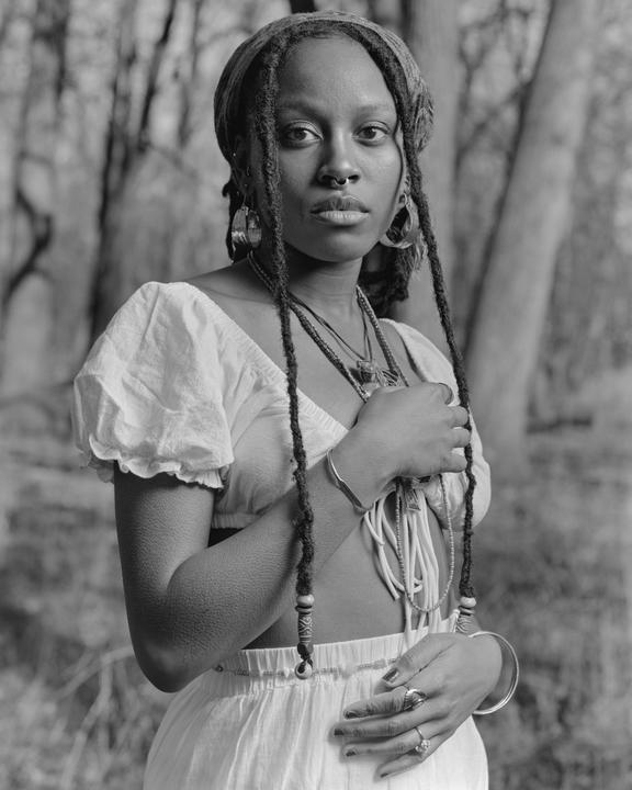 A woman with dreadlocks and a nose ring stands in a forest.