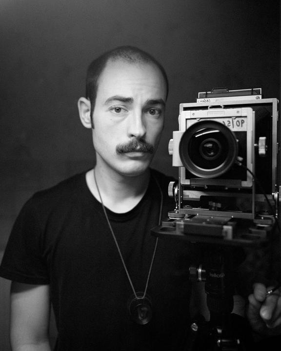 A man with a mustache and a necklace looks at the viewer next to a large format camera.