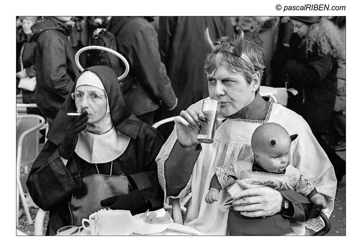 A woman dressed as a nun with a halo smokes a cigarette next to a man dressed as a devil holding a doll with horns and a third eye.