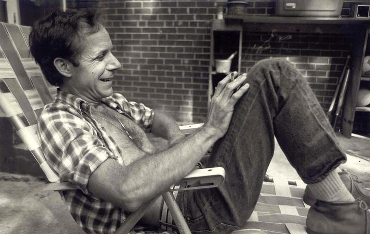 A man in a plaid shirt and jeans sits in a folding chair, smiling and looking to the right.