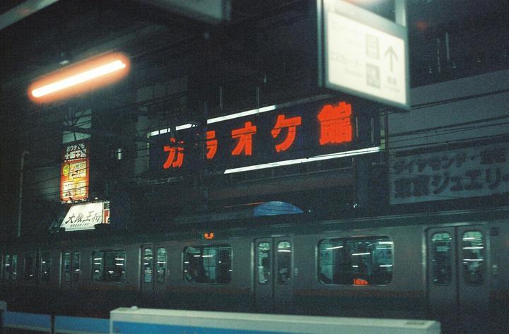 A train is stopped at a station platform at night, with neon signs visible above it.