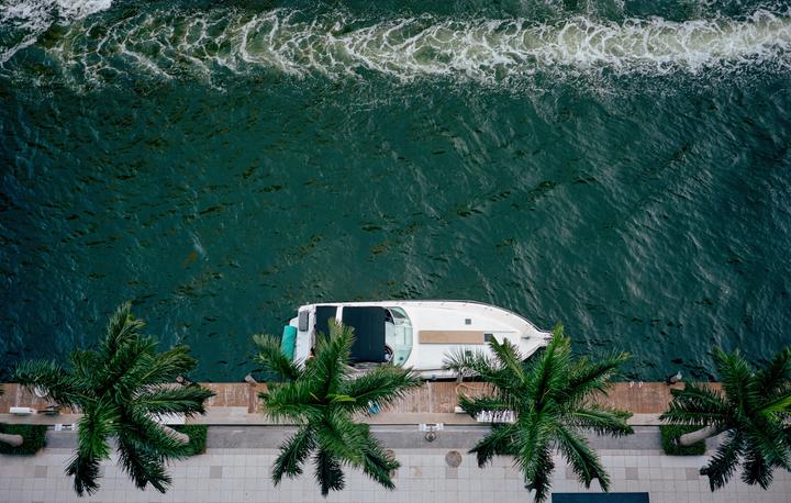 A white boat is docked next to a boardwalk with palm trees, seen from an aerial perspective.
