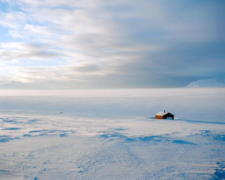 A small, dark cabin sits alone in a vast, snow-covered landscape under a cloudy sky.