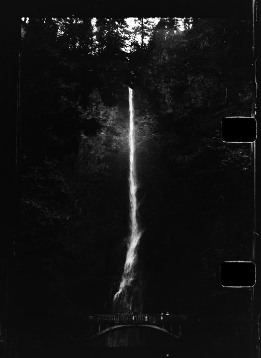 A tall waterfall cascades down a dark, rocky cliff face, with a bridge and people visible at its base.