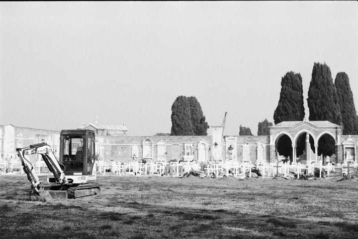 A small excavator sits in a grassy field in front of a cemetery wall with cypress trees and an arched structure.