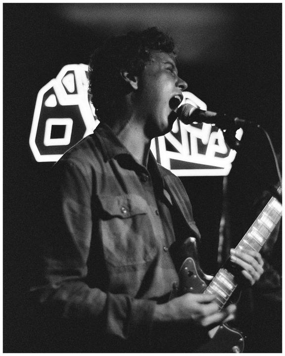 A man sings into a microphone while playing a guitar on a stage with a neon sign in the background.