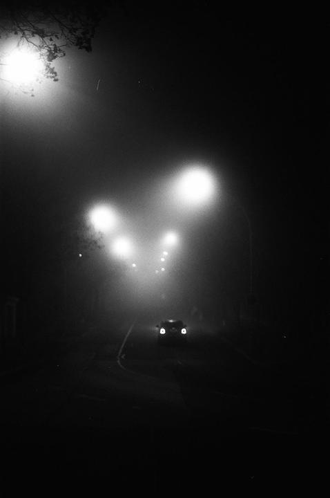 A car with its headlights on drives down a street at night, illuminated by streetlights in the fog.