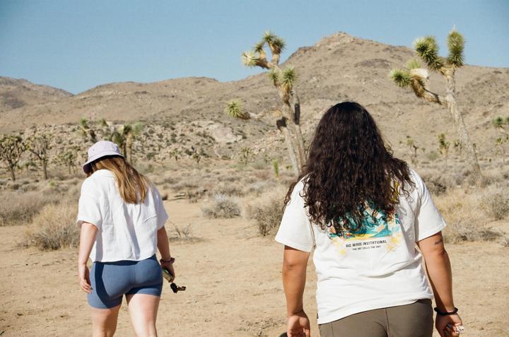 Two women walk away from the camera through a desert landscape with Joshua trees and mountains.