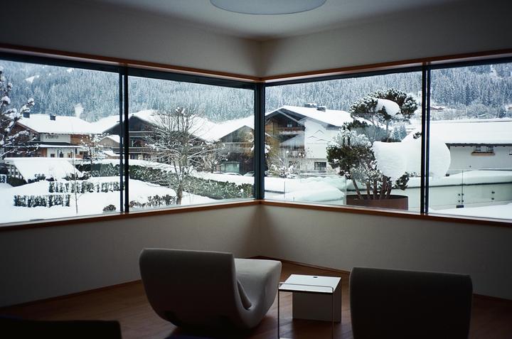 A modern room with two chairs and a small table looks out through large windows onto a snow-covered village and forest.