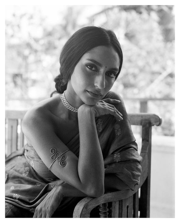 A woman wearing a sari and jewelry sits on a wooden chair, looking towards the viewer.