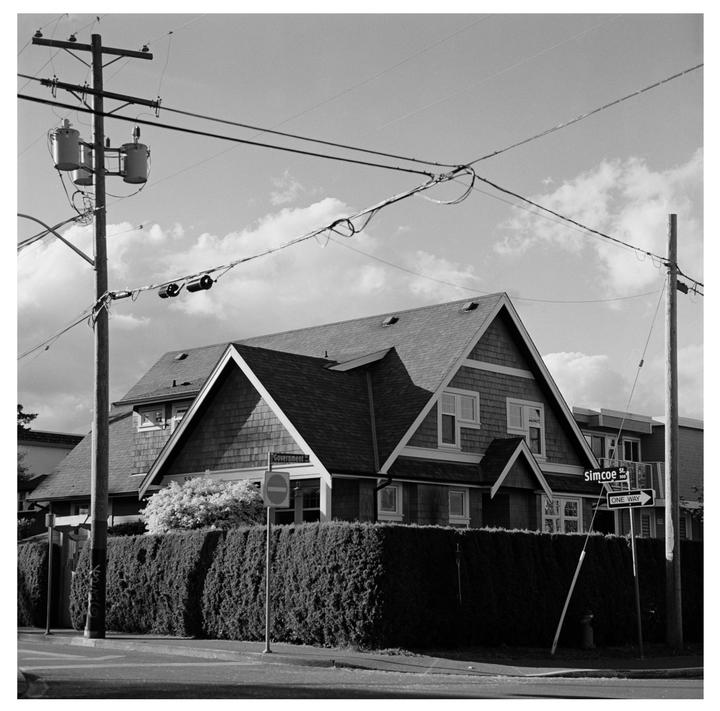 A black and white street photograph shows a house behind a tall hedge, with power lines and utility poles in the foreground.