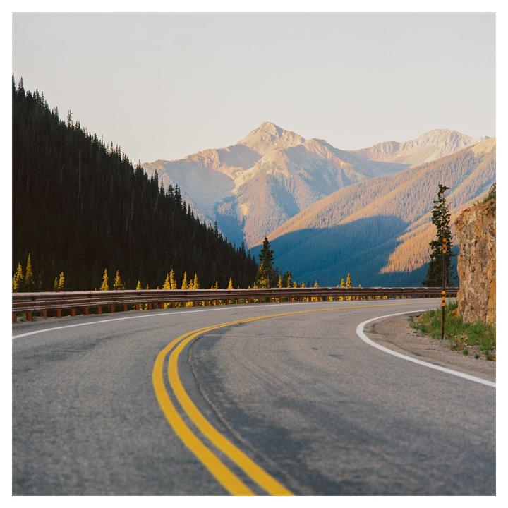 A winding road with a guardrail curves through a mountain landscape with pine trees under a clear sky.