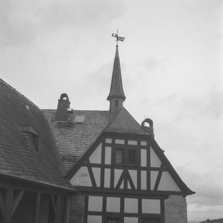 A black and white photo shows the roof and upper facade of a half-timbered building with a spire and weather vane.