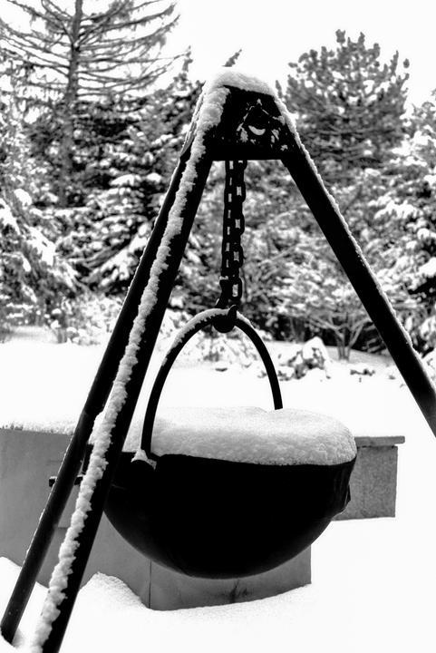 A snow-covered cauldron hangs from a tripod in a snowy forest.