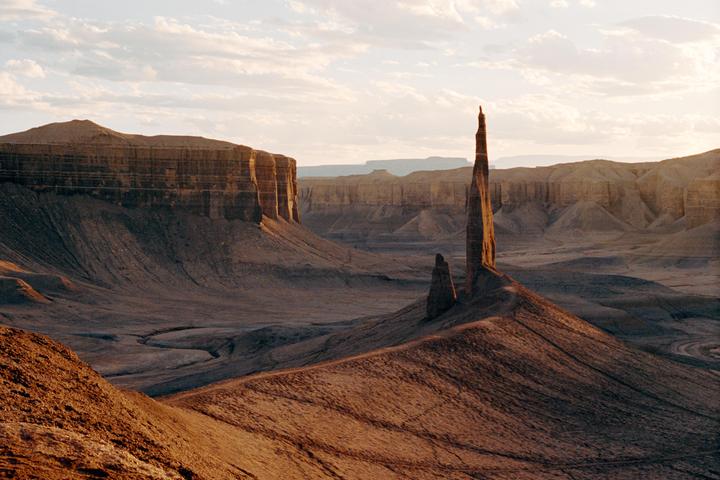 A tall, thin rock spire stands prominently in a vast desert landscape under a cloudy sky.