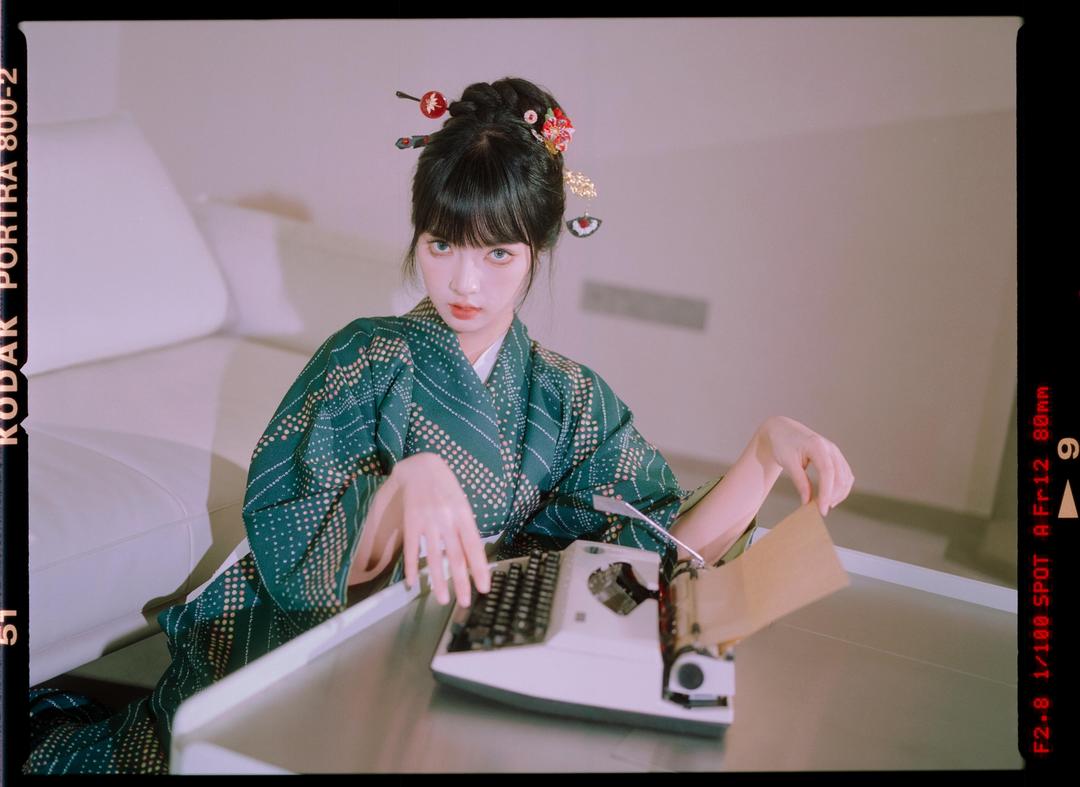A woman in a green kimono with hair ornaments sits at a table with a typewriter.