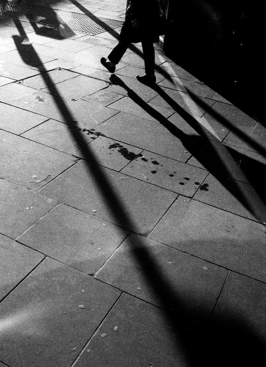 A person walks on a tiled sidewalk with long shadows cast across the ground.