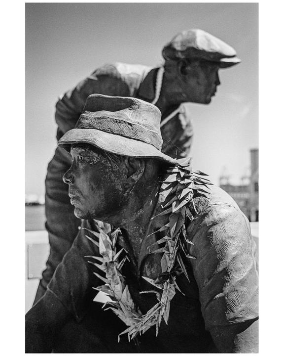Two statues of men, one wearing a fedora and a lei of paper cranes, are shown in profile.