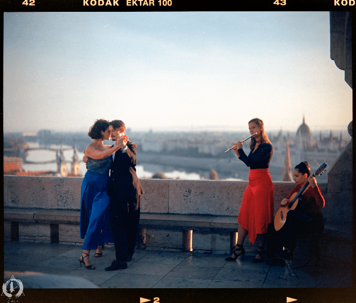 A couple dances on a rooftop while two women play music with a flute and a guitar, overlooking a city.
